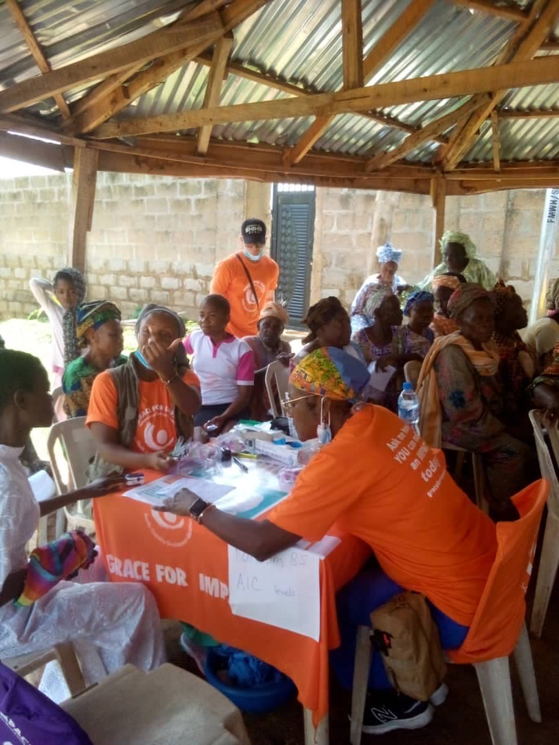 A full clinic in session — community members, mostly elders, seated around the room while volunteers run blood pressure and A1C screenings at the front table.